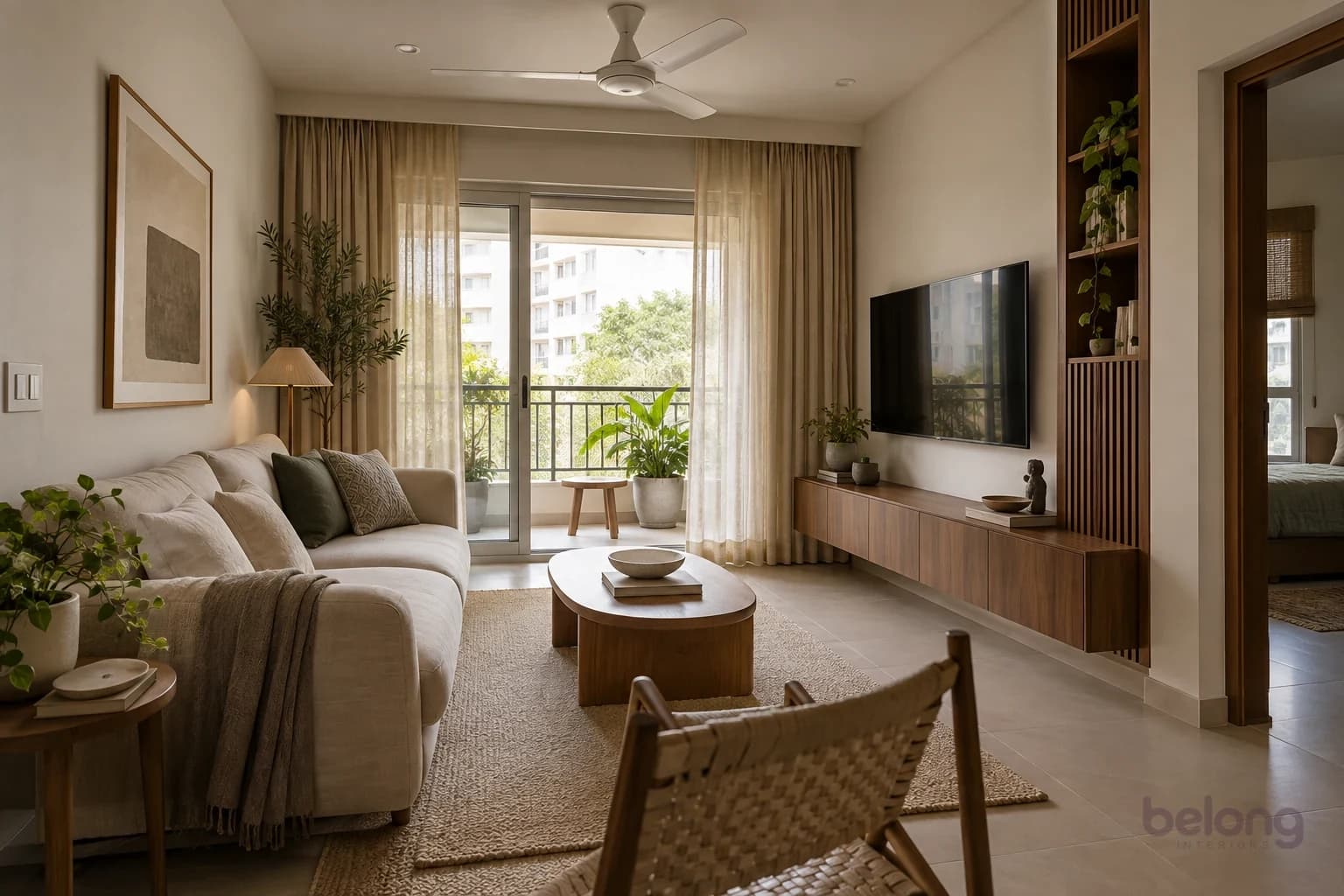A compact living room with a low oatmeal linen sofa, a curved solid-oak coffee table, a slatted walnut storage niche, plants by the balcony door and a white ceiling fan.
