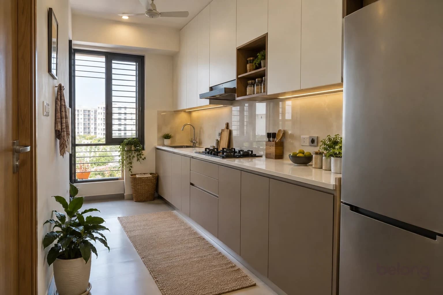 A compact single-wall kitchen with warm grey handle-less base cabinets, ivory ceiling-height upper cabinets, one small open shelf and a mostly clear pale worktop beside a slim fridge.