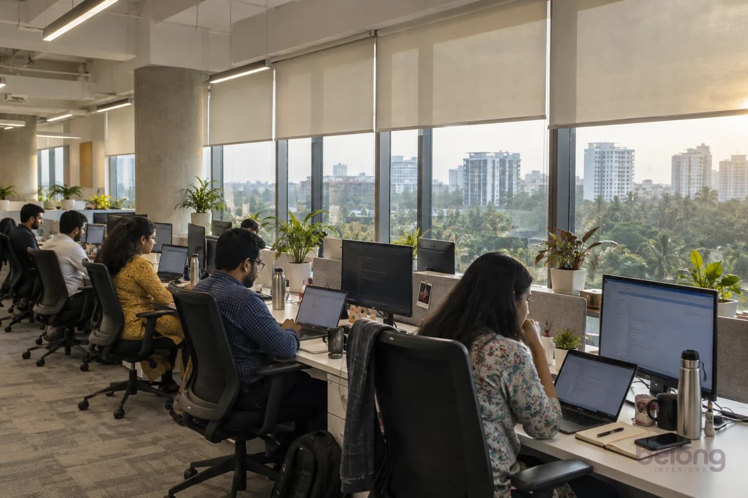 Desks beside large windows with screens at right angles to the glass and roller blinds controlling the sun.