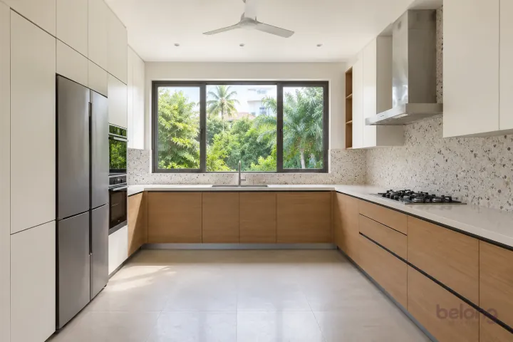 A U-shaped Indian villa kitchen with white cabinetry, oak drawers and a broad central window.