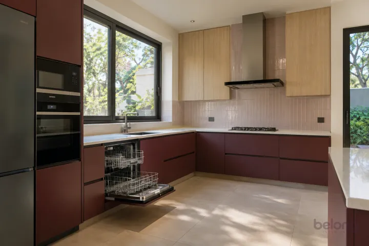 New modern fitted dishwasher with empty racks in burgundy and pale oak, with warm-white solid surface worktops and pale blush rectangular tiles in stacked rows.